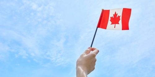 A person’s hand holding a small Canadian maple leaf flag against a bright blue sky with light clouds.