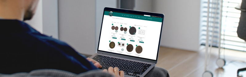 A person holding a smartphone displaying a price chart and gold coin data, with physical gold and silver coins scattered on a wooden table in the background.