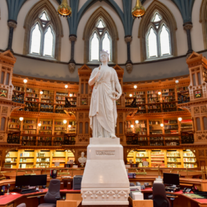 Statue in the center of a grand library reading room with tall arched windows and ornate wooden bookshelves behind it.