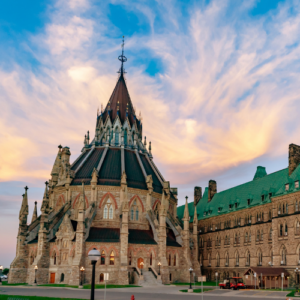 Gothic-style stone cathedral with a tall central spire and ornate buttresses, adjacent brick building, under a colorful sunset sky